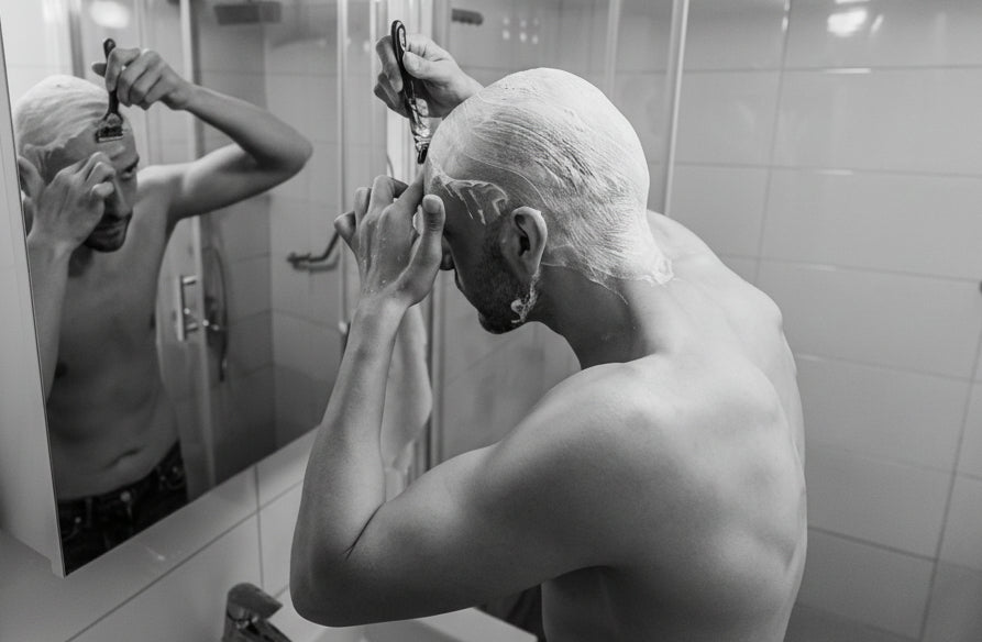 Man shaving his head in front of a bathroom mirror.